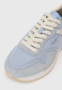 Απαλή μπλε αθλητικά παπούτσια με σουέτ και υφασμάτινα υλικά, που διαθέτουν κρέμα κορδόνια, αντιθετικά σχέδια και μπεζ σόλα. Το λογότυπο της μάρκας ορατό στο πλάι.