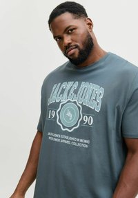 T-shirt in cotone grigio con una grande grafica frontale che presenta il testo "JACK&JONES", un logo circolare e un anno vintage. Maniche corte, design a girocollo.