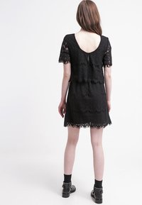 Svart spetsklänning med korta ärmar, öppen rygg, scalloped nederkant och fransdetaljer. Har en figurnära kropp med en texturerad blommönster.