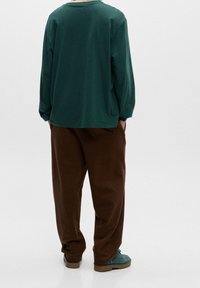 Langærmet, mørkegrøn bomulds skjorte, båret med løse, brune bukser og grønne sko, set fra bagsiden. Glat tekstur, enkelt design.