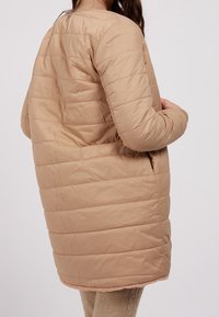 Kvinde iført en lang, polstret beige frakke med horisontal quiltning, stående med den ene hånd i lommen mod en ensfarvet baggrund.