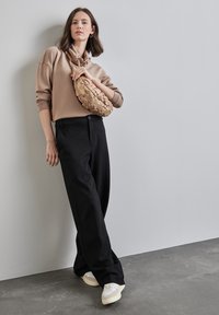 Felpa con cappuccio beige abbinata a pantaloni neri a gamba larga e sneaker bianche. Una borsa tessuta beige è indossata a spalla. Sfondo pulito.