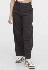 Femme portant un pantalon large noir et des baskets blanches et vert olive, debout devant un fond clair uni.