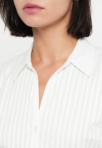 Λευκό ριγέ πουκάμισο με γιακά με κουμπιά. Το υλικό φαίνεται ελαφρύ, με λεία υφή και διακριτική γυαλάδα. Απλό μπροστινό σχέδιο.