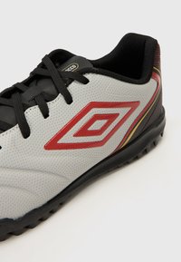 Biele syntetické futbalové topánky s čiernymi šnúrkami, s červeným diamantovým logom a textúrovaným povrchom. Gumová podrážka s malými kolíkmi pre lepšiu trakciu.