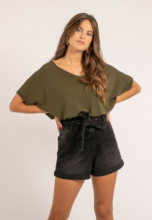 Jeune femme aux longs cheveux ondulés porte un haut ample vert olive et un short en denim noir taille haute avec une ceinture nouée, debout les mains sur les hanches.