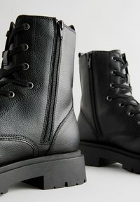 Las botas negras de imitación cuero hasta el tobillo presentan un frente con cordones y una cremallera lateral, superficie texturizada, suela gruesa y un diseño de huella resistente.