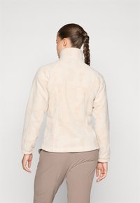 Chaqueta de forro polar en beige claro con un sutil patrón de olas, cuello alto y mangas largas. Fabricada con un material suave para proporcionar calidez y comodidad.