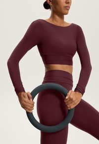 Bordový crop top s dlhým rukávom s hladkou textúrou, skombinovaný s nohavicami s vysokým pásom. Osoba drží okrúhly šedý fitness doplnok.