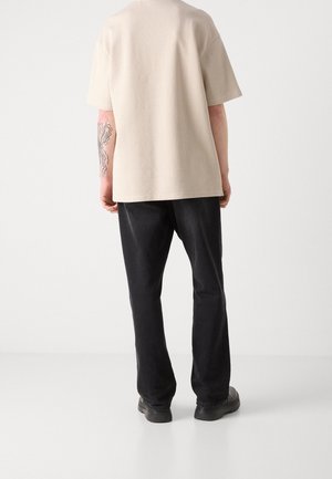 Person with tattooed left arm wearing beige short-sleeve shirt, black pants, and black shoes, standing facing away on white background.