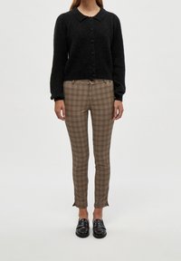 Kvinna iklädd en svart knappad cardigan, bruna rutiga slim-fit byxor och svarta loafers, stående mot en enkel ljus bakgrund.