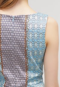 Bezrukávý oděv z modré a fialové vzorované látky. Obsahuje složité geometrické vzory a kontrastní část s dekorativními akcenty.