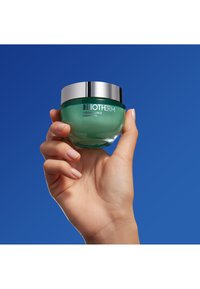 Crema notte Biotherm Aquasource in un barattolo di vetro trasparente con un coperchio argentato; crema verde chiaro visibile all'interno; tenuta in una mano contro uno sfondo blu.