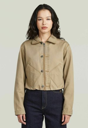 Donna con lunghi capelli scuri che indossa una giacca beige con zip e tasche anteriori inclinate e jeans scuri a vita alta, in piedi davanti a uno sfondo semplice.