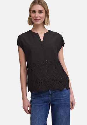 Frau trägt eine schwarze Kurzarmbluse mit bestickten Cut-out-Mustern, kombiniert mit blauen Jeans, steht vor einem neutralen Hintergrund.