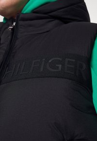 Zbliżenie na osobę w czarnej ocieplanej kamizelce z napisem "HILFIGER" na klatce piersiowej, noszącą zieloną bluzę z kapturem.