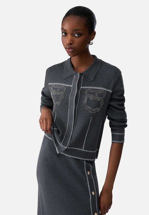 Donna che indossa un completo top e gonna in maglia grigio scuro con dettagli di cuciture bianche e bottoni dorati sul lato della gonna, posando con la mano sul fianco.