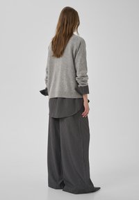 Grauer Strickpullover über einem schwarz-weiß gestreiften Hemd und weit geschnittenen Hosen. Weiche Textur, lockere Passform, hochgekrempelte Ärmelabschlüsse.