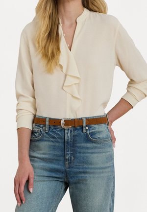 Frau trägt eine cremefarbene Bluse mit langen Ärmeln und Rüschen-Detaillierung, in eine hochgeschnittene blaue Jeans mit braunem Ledergürtel gesteckt.