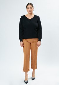 Schwarzer V-Ausschnitt-Pullover mit Glitzer, kombiniert mit beige-gefärbten, taillierten Hosen, die an den Knöcheln gekrempelt sind. Schwarze, spitze Absätze mit floralem Schmuck.