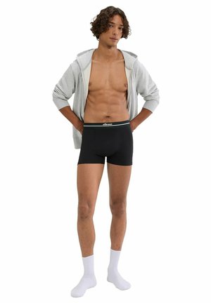 Young man wearing gray zip-up hoodie, black boxer briefs with waistband logo, and white socks, standing with hands on hips.