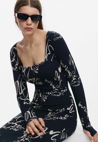 Pikad varrukad, tumehall bodysuit kreemja abstraktse käsikirjaga mustriga. Omab ruudukujulist kaelust ja ribakujulist tekstuuri. Mustad prillid kuuluvad komplekti.