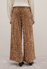 Weite Hose aus goldfarbenem Paillettenstoff mit einer strukturierten Oberfläche und schimmerndem Effekt. Die Schuhe sind unter dem Saum sichtbar.