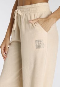Beige Lounge-Hosen aus weichem Stoff, mit einem elastischen Taillenbund und innerem Kordelzug sowie Seitentaschen. "FEEL GOOD" in silbernen Akzenten.