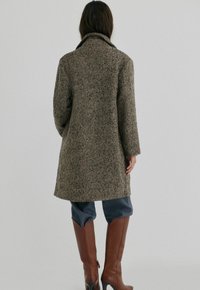 Uldblandet frakke i brun herringbone mønster, med krave og lange ærmer, parret med brune læderstøvler til knæene og blå bukser.