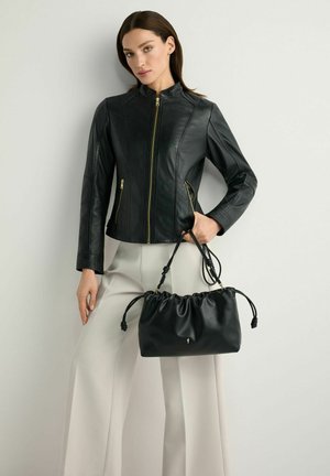 Kobieta ubrana w czarną skórzaną kurtkę zapinaną na zamek i jasne, szerokie spodnie, trzymająca czarną skórzaną torebkę z marszczonym zapięciem za paski.