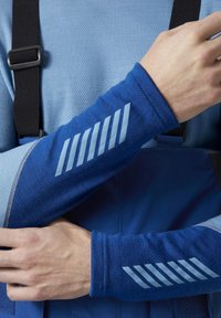 Modrá košile s dlouhým rukávem vyrobená z texturované látky, se tmavě modrými akcenty a světle modrými diagonálními pruhy na rukávu. Viditelné nastavitelná černá poutka.