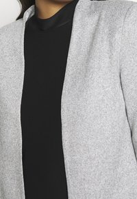 Šedý strukturovaný blazer s otevřeným předním dílem, v kombinaci s černým topem s vysokým límcem. Blazer má volný střih a měkkou látku.