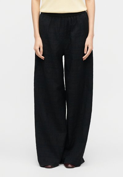 Personne portant un pantalon large noir ample, un haut beige rentré à l'intérieur, et des chaussures marron foncé sur un fond clair uni.