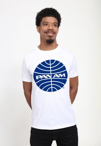T-shirt bianco in cotone con un grande grafico circolare blu con un motivo a griglia e il testo "PAN AM" in lettere grasse al centro.