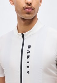 Άνδρας που φοράει λευκό αθλητικό τζέρσεϊ με φερμουάρ και μαύρο κάθετο λογότυπο "OAKLEY", ορατό από τον λαιμό μέχρι το μέσο του στήθους.