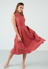 Roze midi haljina s uskim korzetom, lepršavom suknjom i detaljima naboranja. Glatka tkanina i bez rukava, uparen s laganim sandalama na remenčiće.