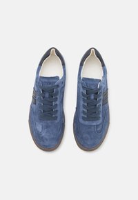 Blå mockaskor med mörka snören, vit innerfoder och en brun gummisula. Har en sidostrikedesign. Platt, rund tå.