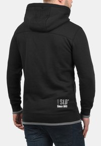 Schwarzer Hoodie mit Kängurutasche, gerippten Bündchen und Saum. Mit einem Logo "Since 1993" auf dem unteren Rücken. Glatter Stoff, lässiges Design.