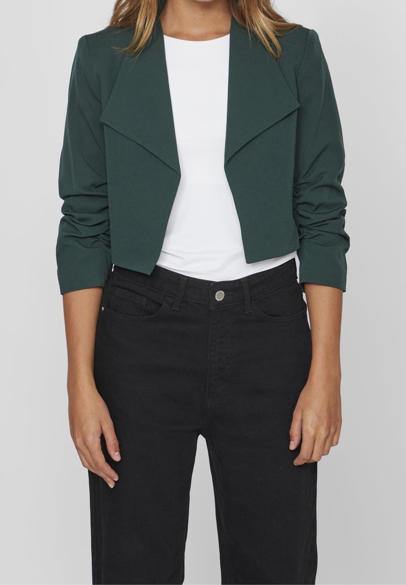Femme portant une veste courte vert foncé sur un top blanc avec un jean noir taille haute, debout les bras détendus.