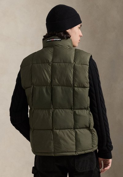 Πράσινο γιλέκο puffer με τετράγωνο σχέδιο ραφής, ψηλό γιακά και λογότυπο στην πλάτη. Φοριέται πάνω από μαύρο μάλλινο πουλόβερ με υφή. Μαύρο σκουφάκι συμπληρώνει την εμφάνιση.