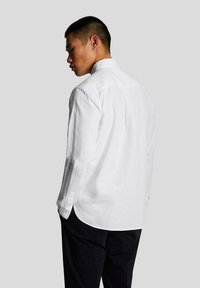 Camisa blanca de manga larga hecha de tejido ligero, con un cuello clásico, puños con botones y un corte recto con un pliegue trasero.