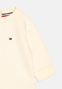 Sweat-shirt couleur crème avec un tissage texturé, des manches raglan et un petit patch logo en bleu marine et rouge sur la poitrine.