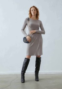 Femme avec des cheveux bouclés portant une robe grise à manches longues en côtes, des bottes noires jusqu'au genou et un petit sac noir avec une chaîne en or.