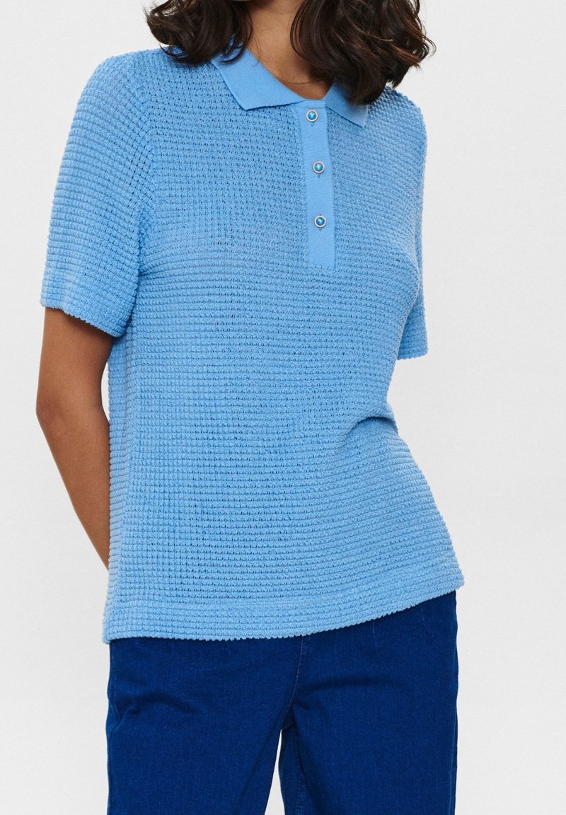 Femme portant un polo bleu clair texturé à manches courtes et un pantalon bleu foncé sur un fond clair uni.