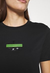 Černé bavlněné tričko s texturovaným černým logem "CK" a zeleným obdélníkovým štítkem, na kterém je bílý nápis "CALVIN KLEIN JEANS".