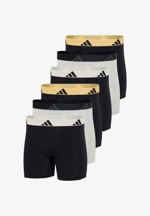 Athletischer Mann steht barfuß vor weißem Hintergrund, trägt schwarze Boxershorts mit einem gelben Bund, der schwarze Logos zeigt.