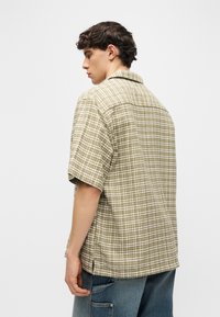 Νέος άνδρας με σκούρα, σγουρά μαλλιά, ντυμένος με πράσινο και άσπρο πουκάμισο με κοντά μανίκια και μπλε τζιν, στραμμένος προς τα αριστερά μπροστά σε λευκό φόντο.