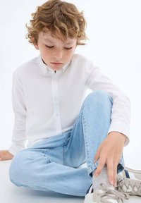 Chemise blanche à manches longues avec boutons, jeans en denim bleu clair, chaussettes grises, et baskets grises/blanches. Enfant assis en tailleur sur un fond blanc.