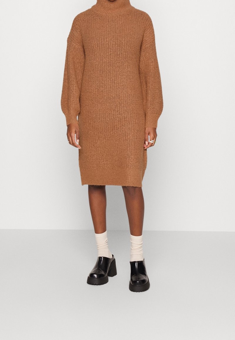 Abito knit marrone con collo alto, maniche lunghe, spacchi laterali e un motivo testurizzato. Abbinato a scarpe con tacco largo nere e calze beige.