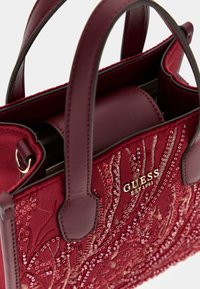 Burgundska torba od teksturirane tkanine s šljokicama, kožnim drškama i zlatnim logotipom. Kompaktni dizajn s strukturiranim oblikom.